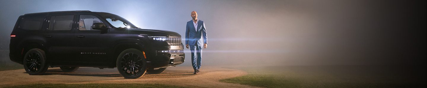 A profile of a 2022 Grand Wagoneer Obsidian with its headlamps on, parked on the pitcher's mound of a baseball field at night with a light beaming from the foggy background. Derek Jeter is walking in front of the vehicle.